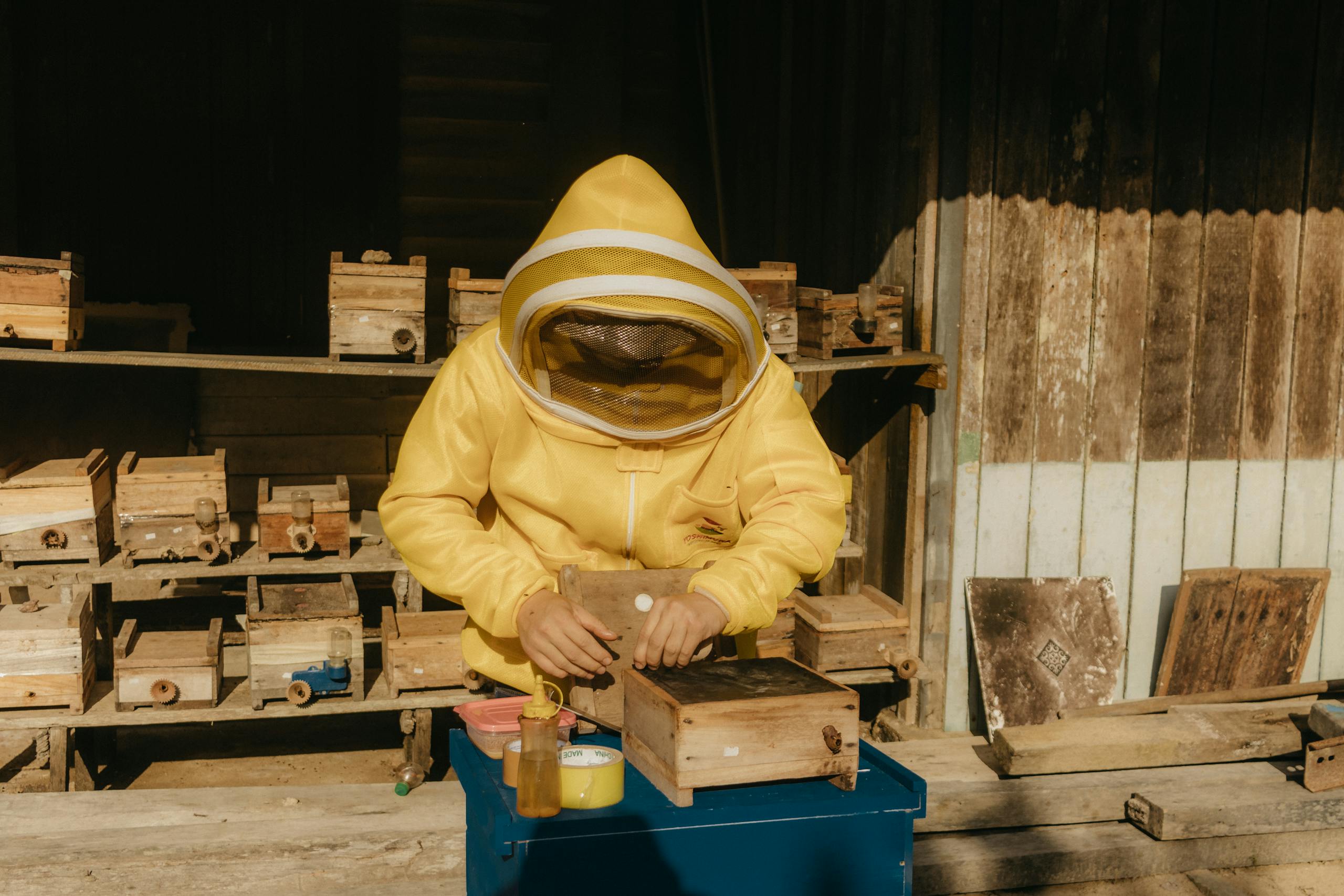A beekeeper in a yellow protective suit works with wooden beehives on a sunny day.